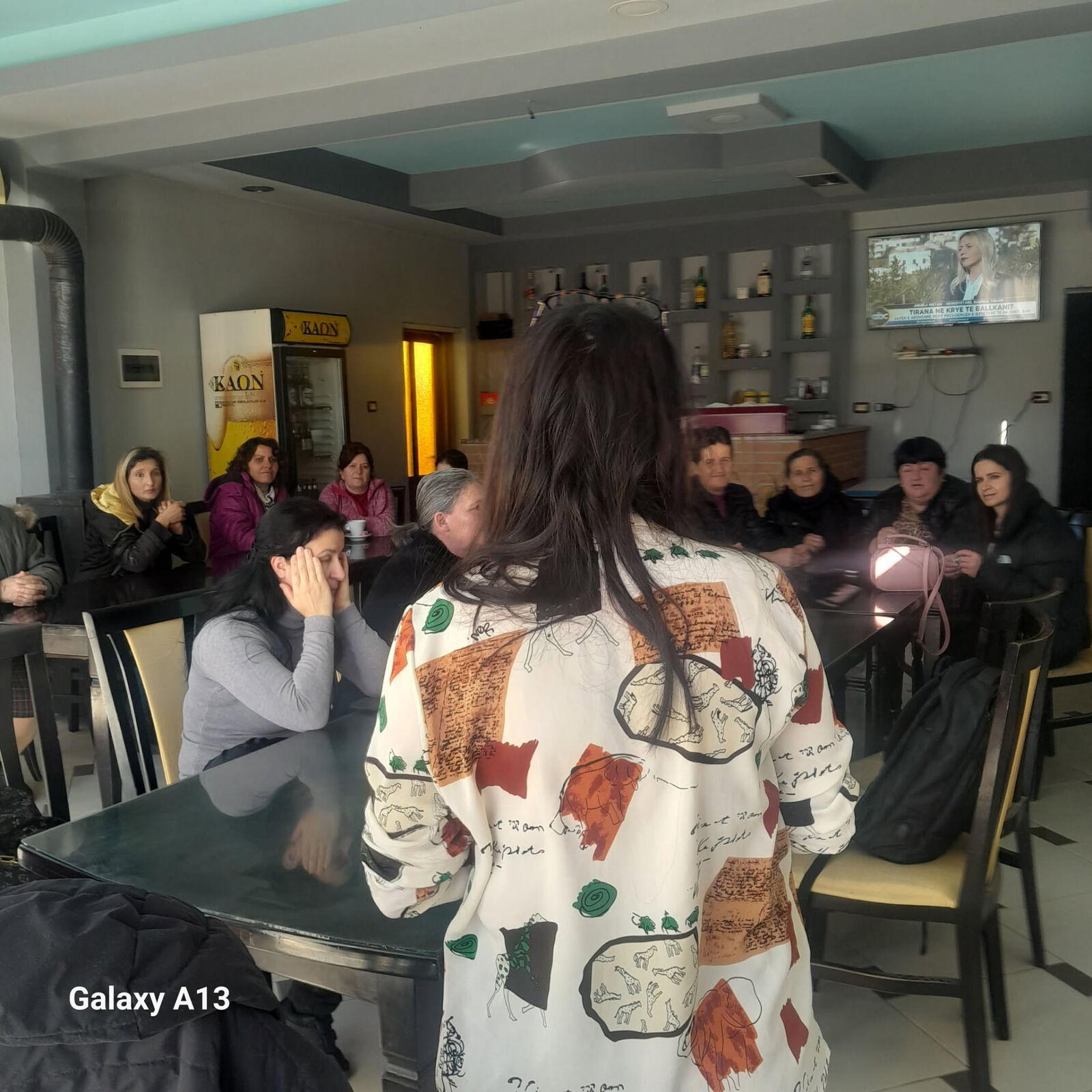 Një grua flet para një grupi grash të ulura rreth tavolinave në një sallë takimesh